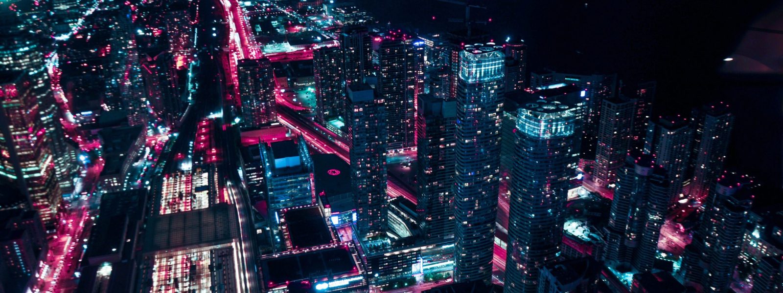 Aerial view of a city at night, illuminated by pink and blue lights, showcasing tall buildings and a winding road with glowing traffic.