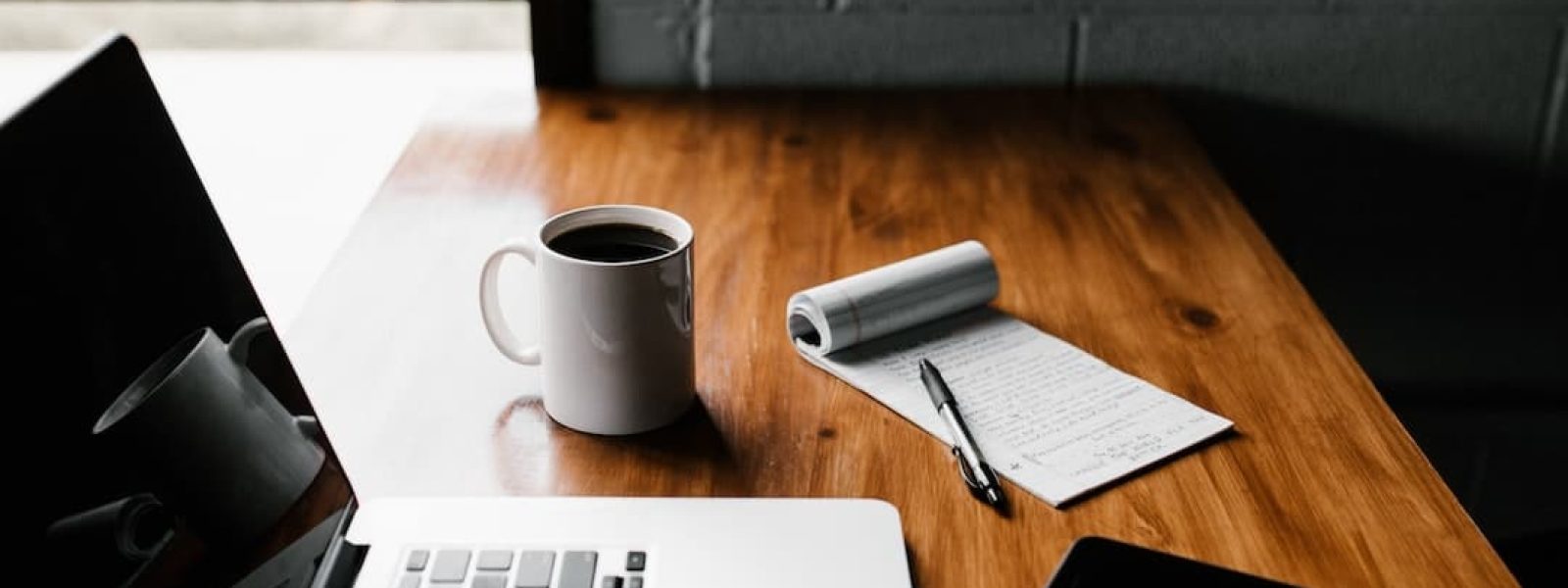 A wooden desk with a laptop, a cup of coffee, a notepad with a pen, and a smartphone.