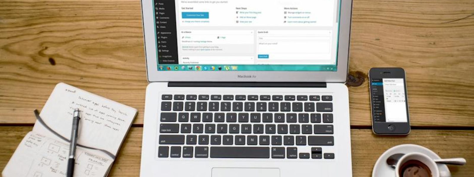 Laptop displaying a website dashboard next to a notepad, smartphone, and cup of coffee on a wooden table.